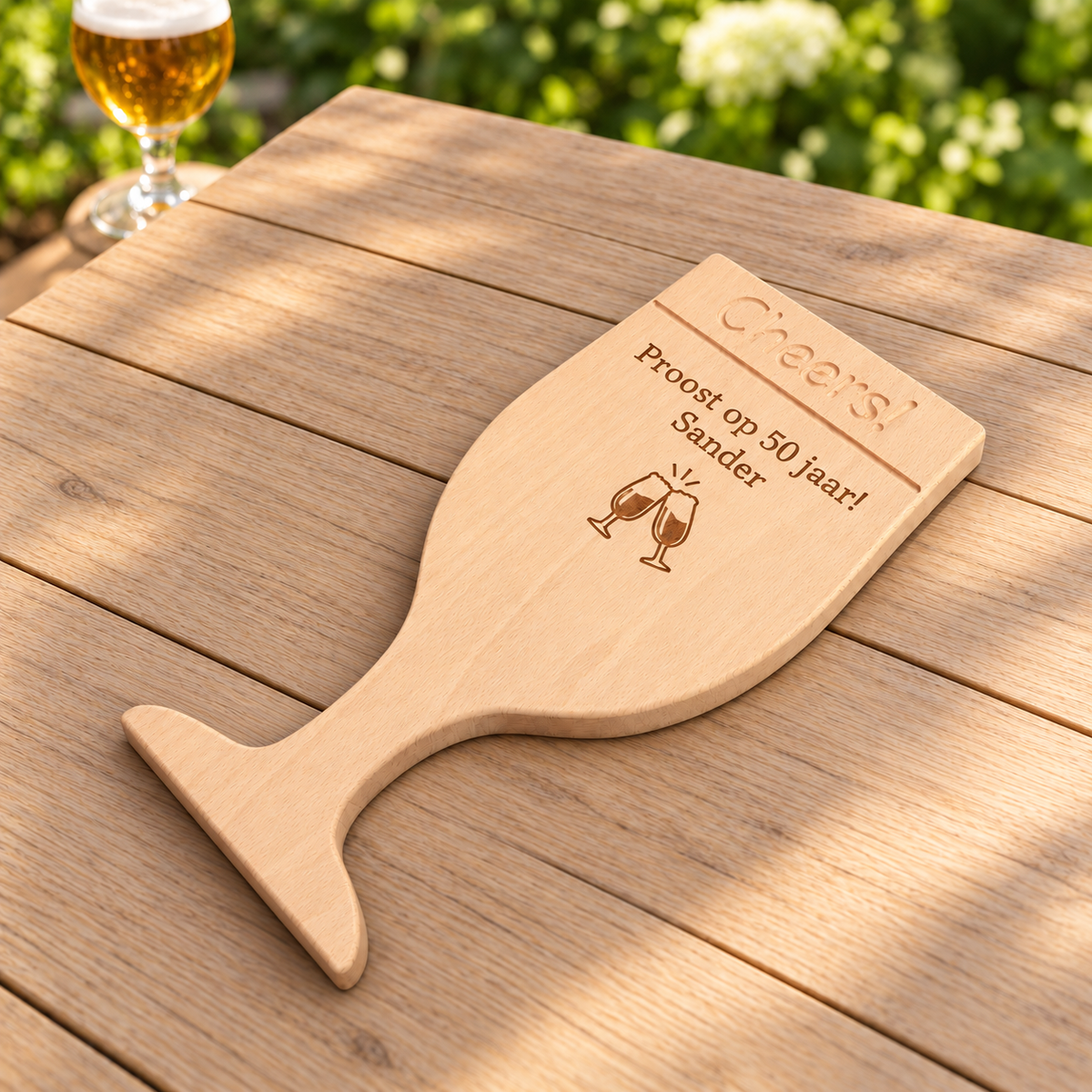 Beukenhouten borrelplank van DecoSpace in de vorm van een speciaalbierglas, met ingefreesd ‘Cheers!’ en de gravure ‘Proost op 50 jaar! – Sander’, op een houten terrastafel.