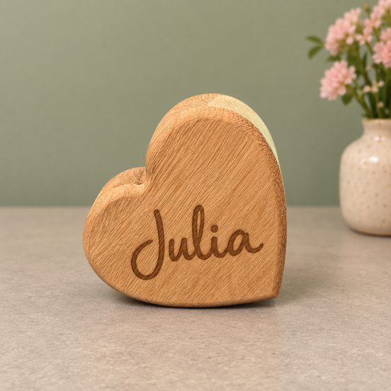 Massief eiken hart van DecoSpace met de naam Julia gegraveerd, op een lichte tafel met roze bloemen op de achtergrond.