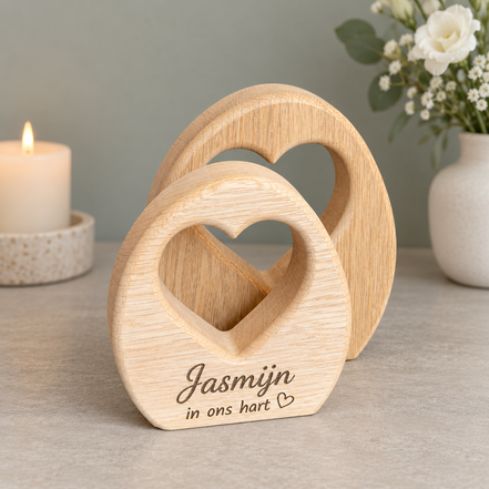 Twee eiken herdenkingsobjecten van DecoSpace met hartvormige uitsparingen, waarvan het voorste gegraveerd is met ‘Jasmijn, in ons hart’, naast een kaars en witte bloemen.