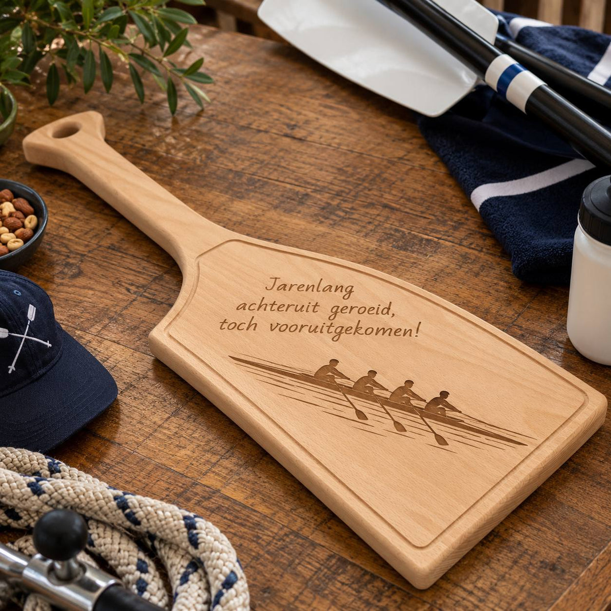 Gepersonaliseerde roeiriem borrelplank van massief beukenhout met gravure van vier roeiers en de tekst ‘Jarenlang achteruit geroeid, toch vooruitgekomen!’, gepresenteerd op een rustieke houten tafel.